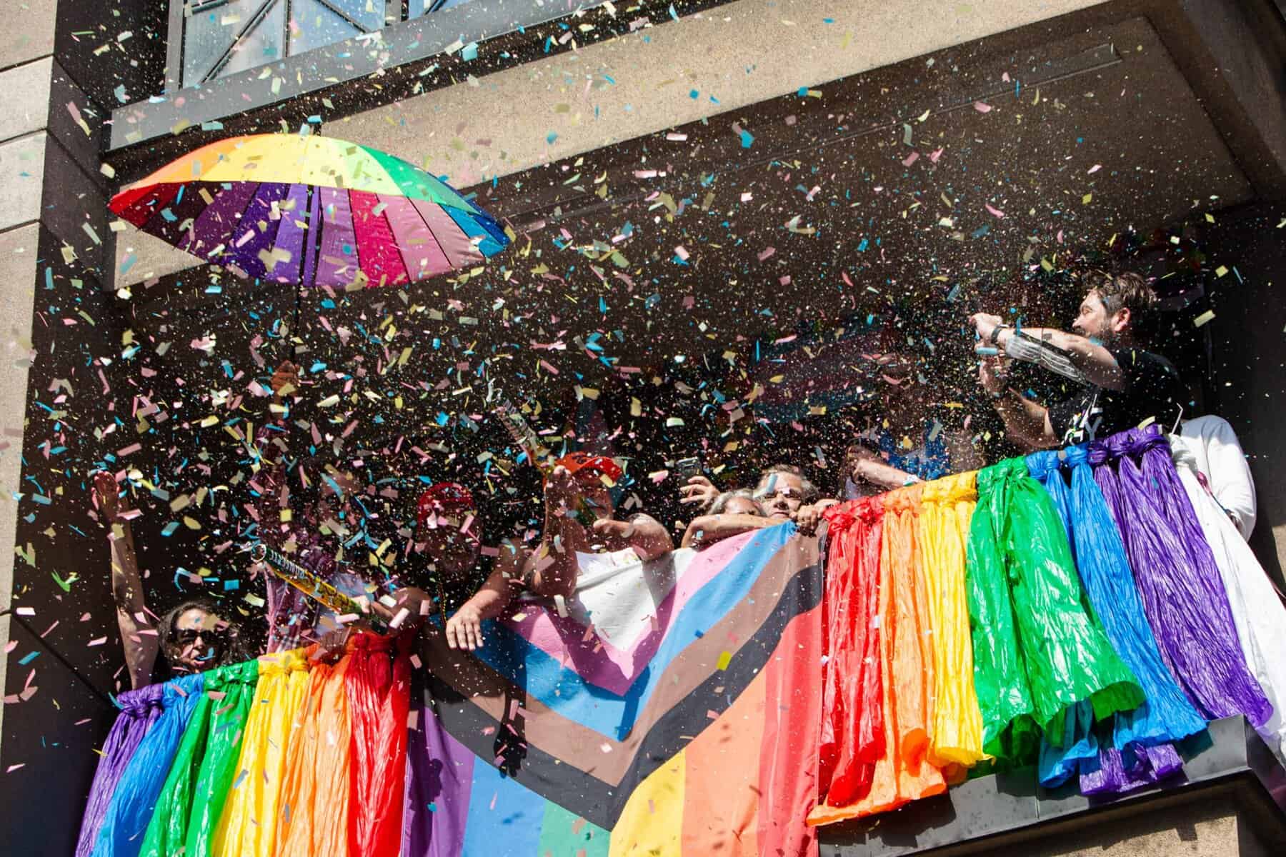 Colorful pride parade celebration with rainbow flags, rainbow umbrella, and confetti on a balcony in Toronto, LGBTQ+ Pride event, inclusive Pride parade, Toronto pride festival, vibrant pride festivities.