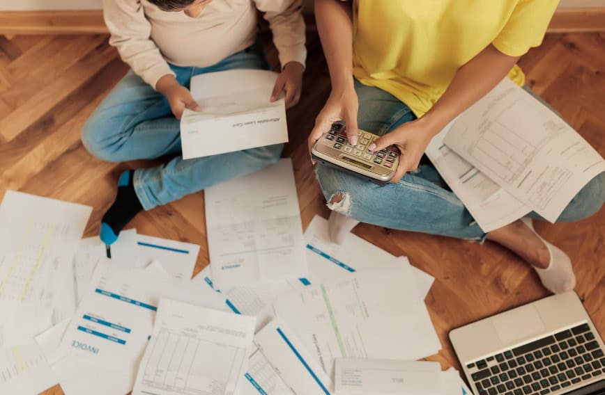 Disorganized papers, calculator, and laptop on wooden floor representing financial planning or budgeting.