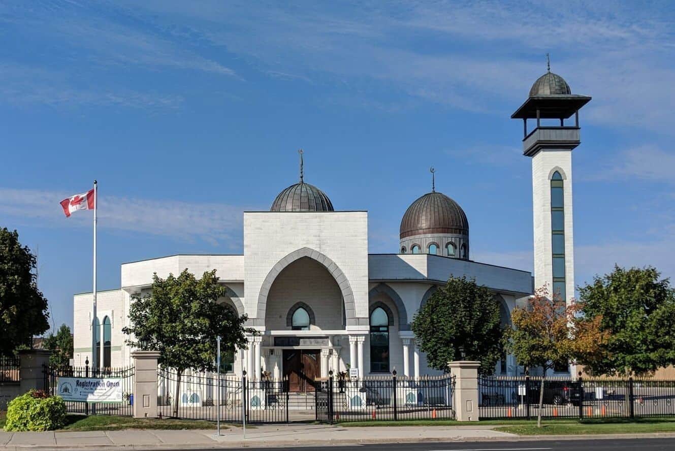 Magnificent white mosque with domes and minaret in Toronto, surrounded by trees and a Canadian flag, showcasing diverse religious architecture in the city.