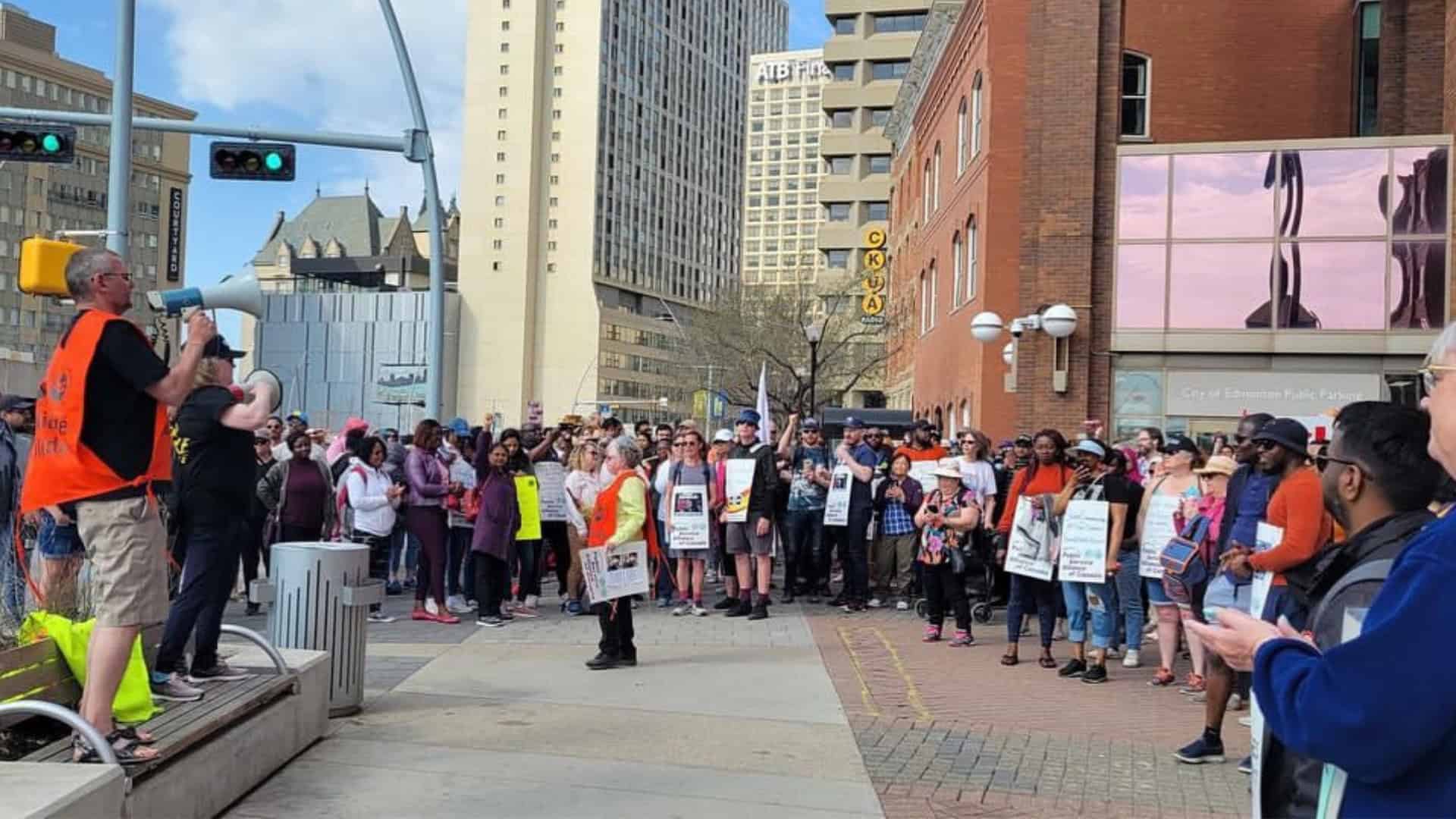 Protest in downtown Toronto with diverse crowd holding signs and listening to speaker, vibrant cityscape backdrop.