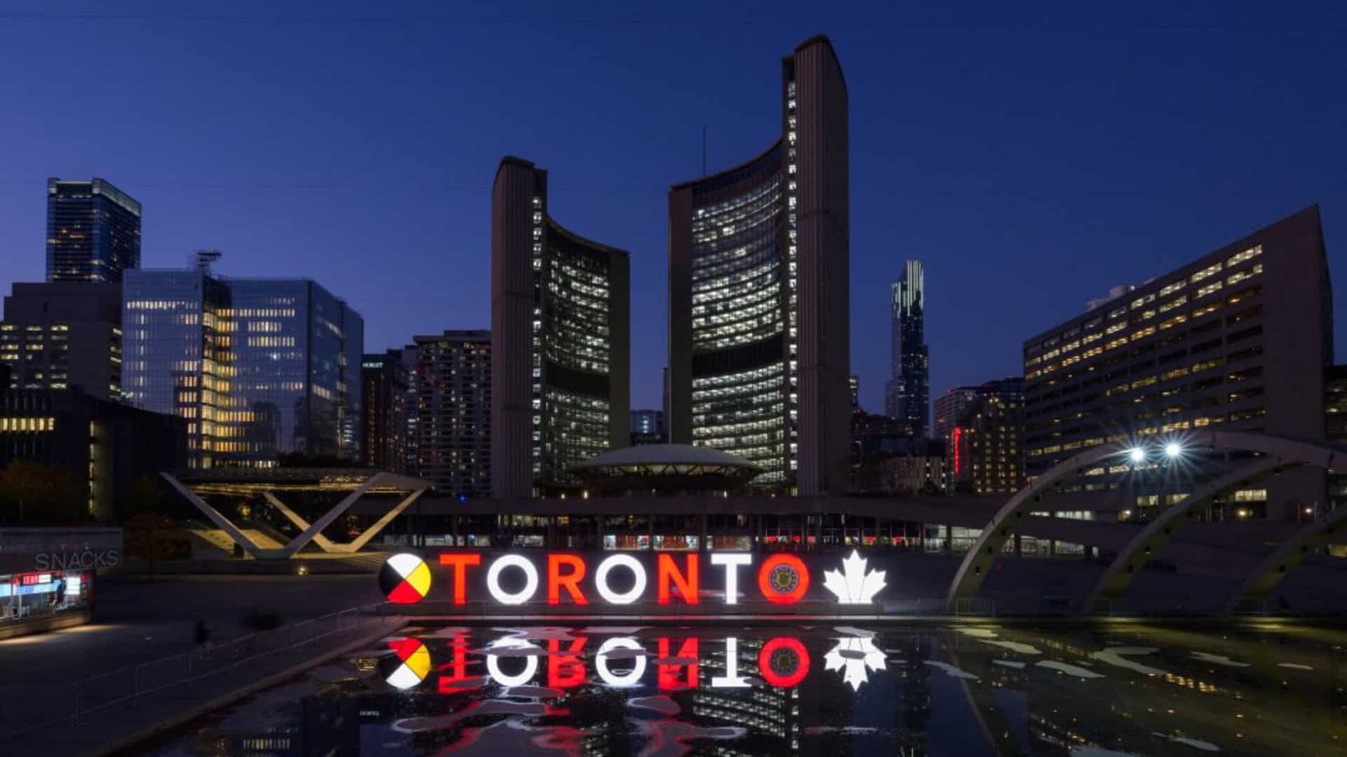 Night view of Toronto city skyline with illuminated buildings and Toronto sign reflected in the water, showcasing vibrant city life and architecture.