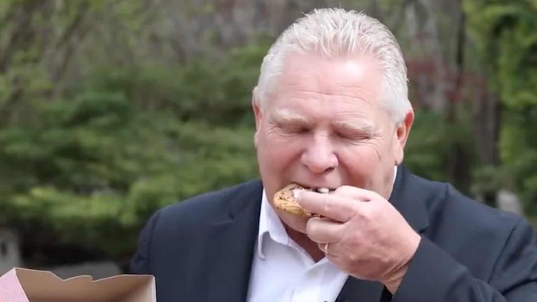 Scruffy man eating cookie outdoors in the park, dressed in a business suit, enjoying a casual snack while smiling.