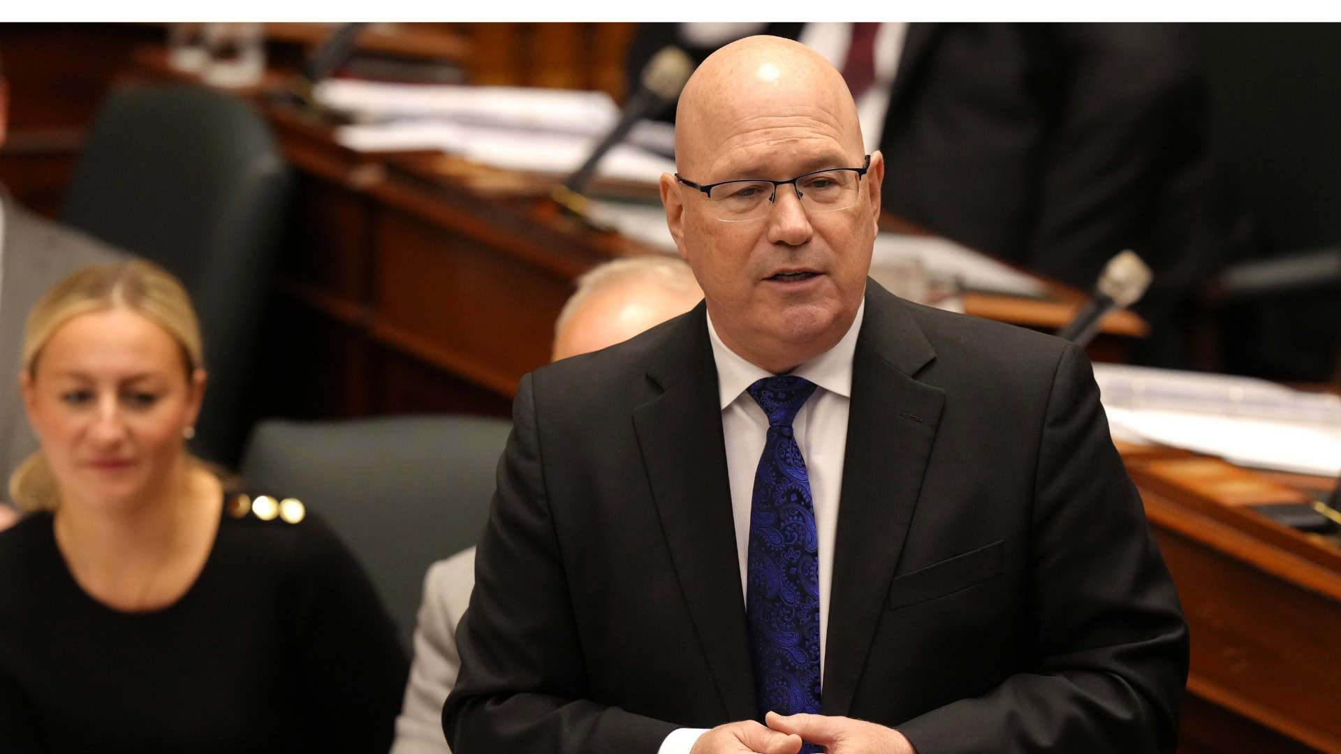 Bald man in suit speaking in a parliamentary setting with colleagues around him.