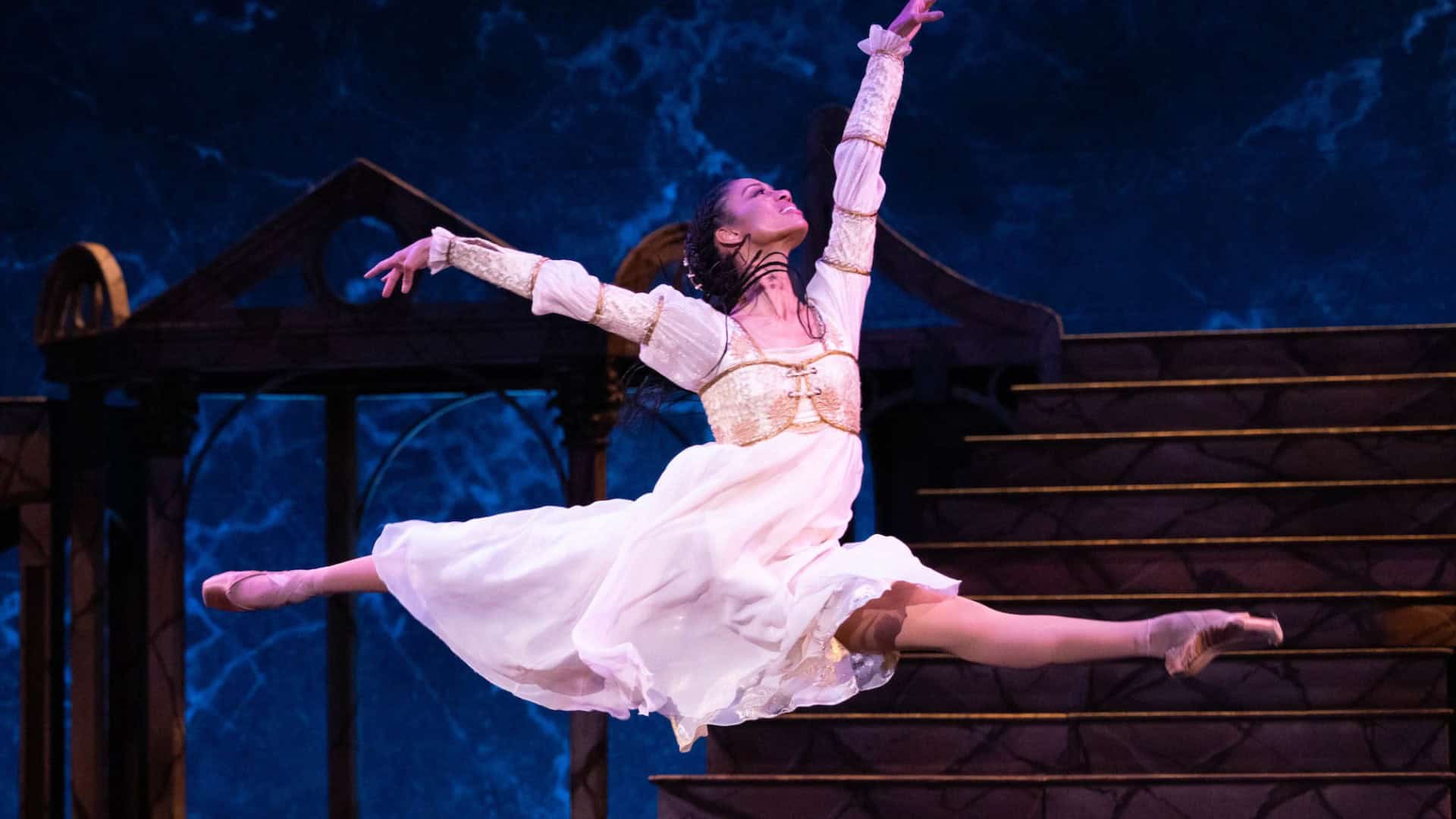 Elegant ballet dancer performing on stage in a flowing white costume with a dark blue background, showcasing Toronto arts and culture in a theatrical dance production.