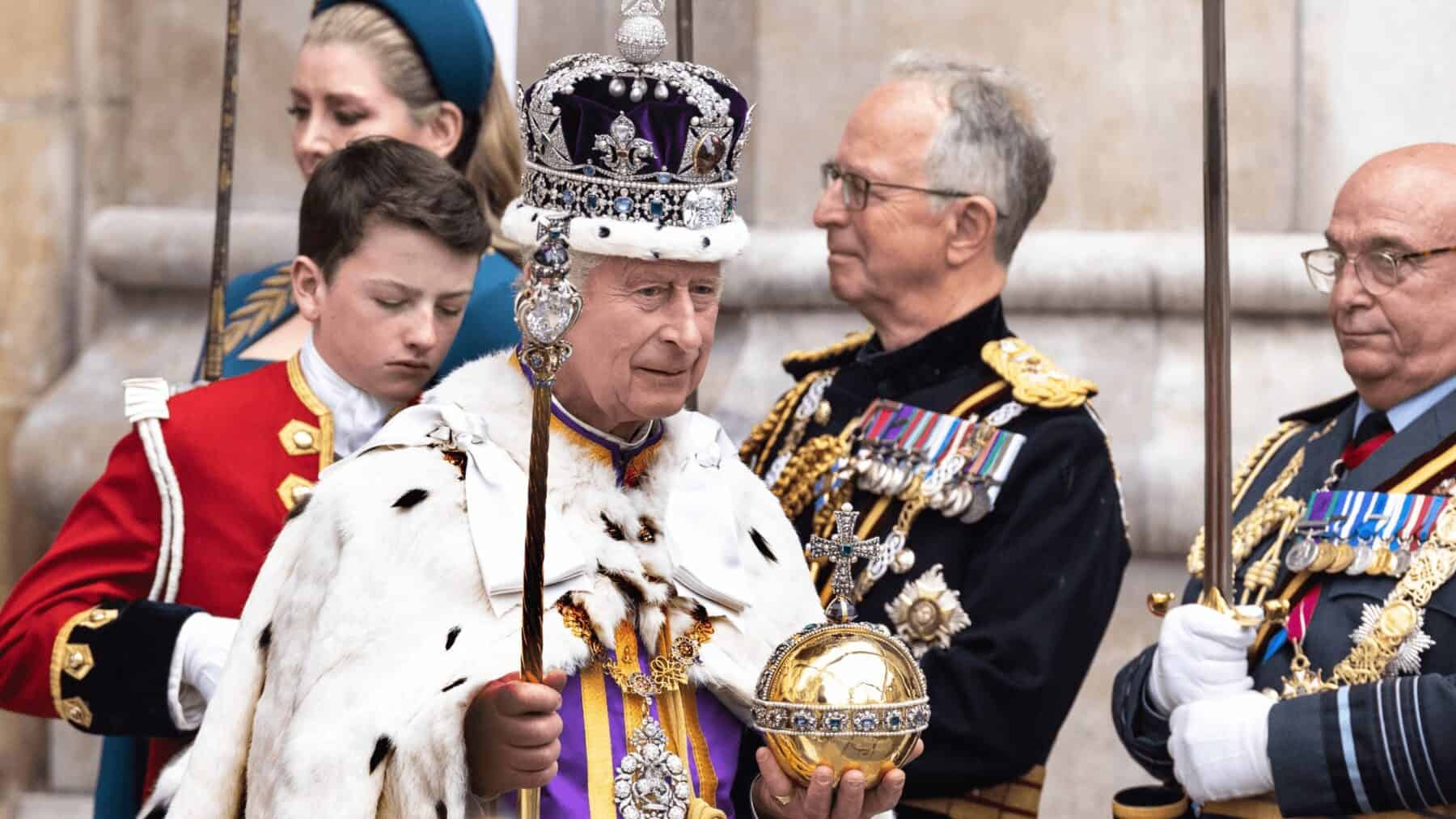 King Charles III wearing a crown and robes, participating in a ceremonial event with military personnel and attendants. Traditional royal event at a historic venue.