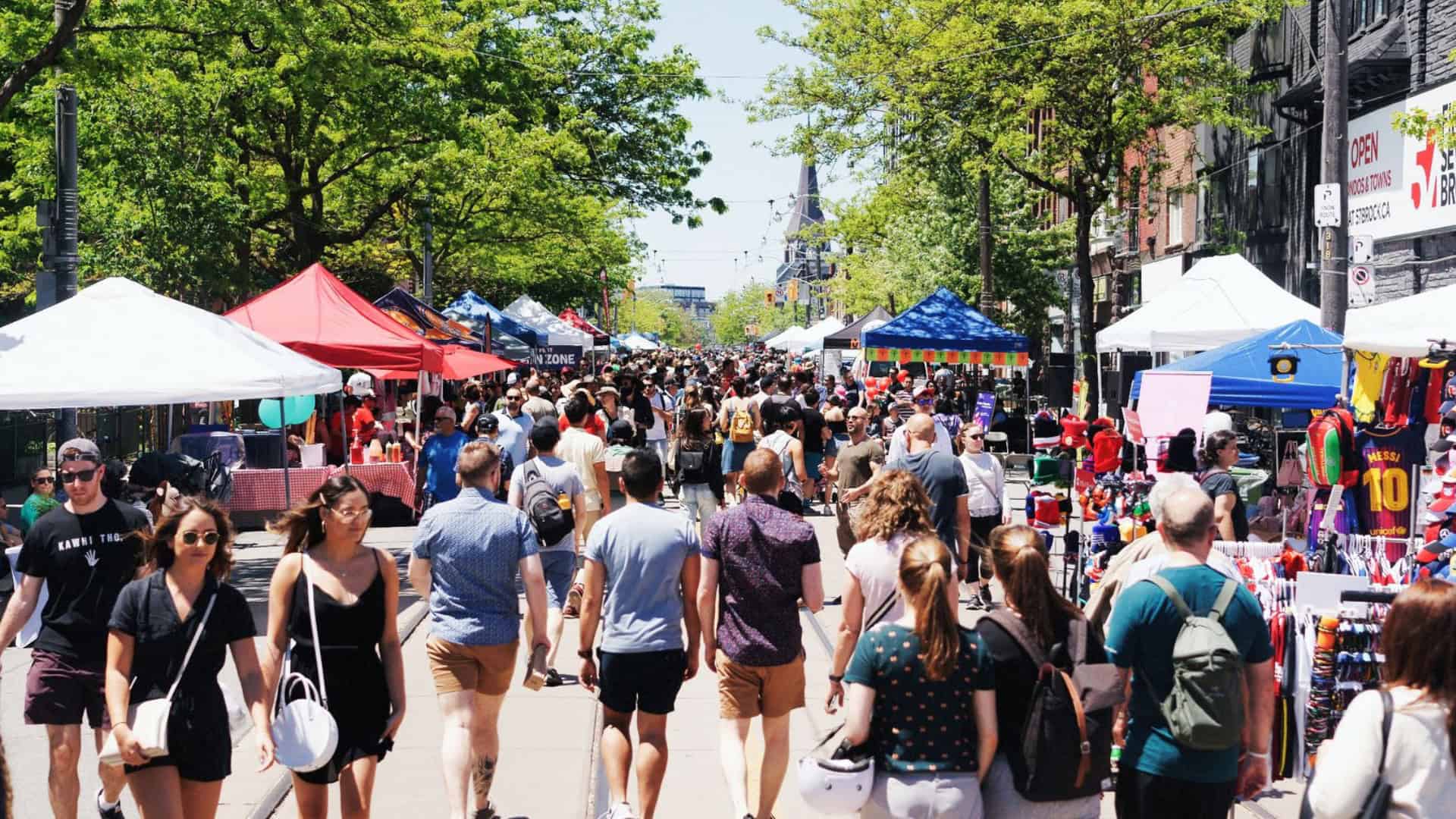 Colourful outdoor street market in downtown Toronto during a lively festival, with vendors and crowds enjoying local arts, crafts, food, and entertainment on a sunny day.