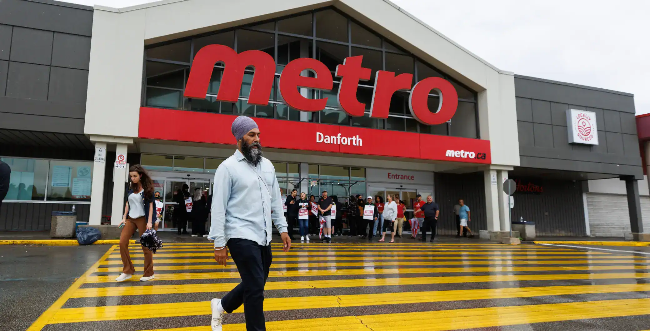 Metro Toronto store exterior, shoppers entering Danforth location, Toronto retail, public transit hub, urban shopping district, street crosswalk, rainy weather Canada, community shopping scene, NOW Toronto.