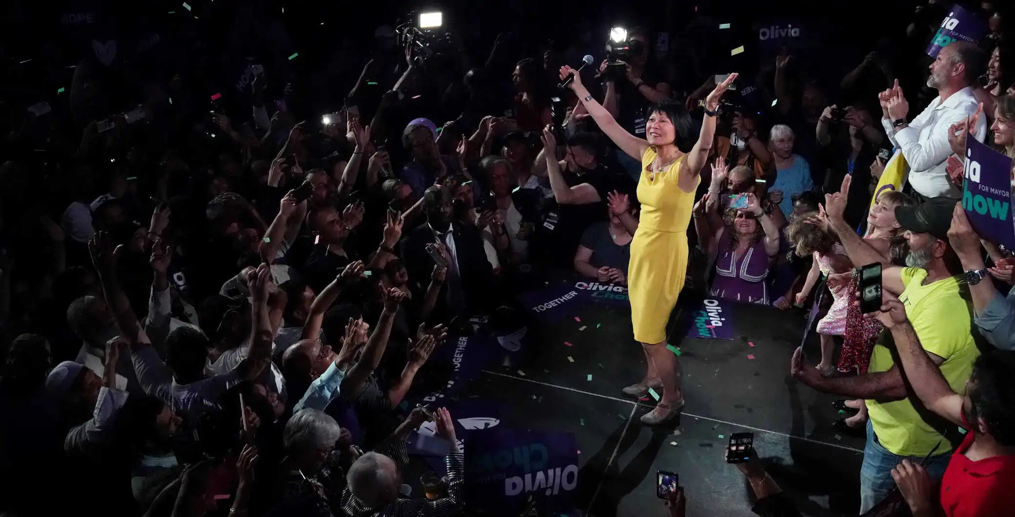 Vibrant crowd cheering and celebrating at a political campaign event in Toronto, with Olivia Chow in a yellow dress engaging with supporters.