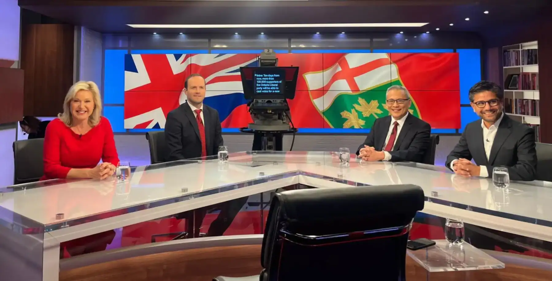 Diverse panelists seated at a modern conference table during a televised discussion, with flags of Canada and the United Kingdom in the background, representing news and current events in Toronto.