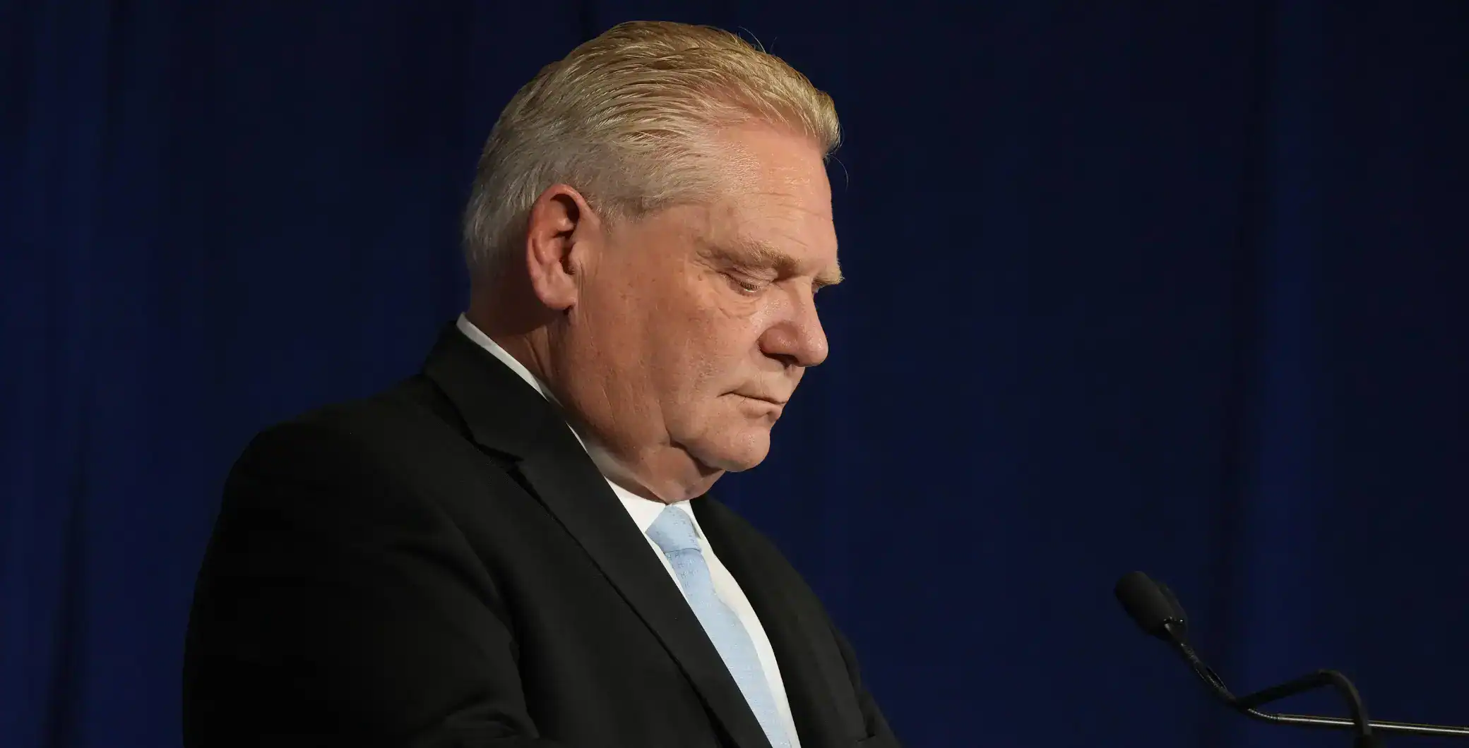Senior man in formal suit with hair combed back looking down, during a speech or presentation at an event.