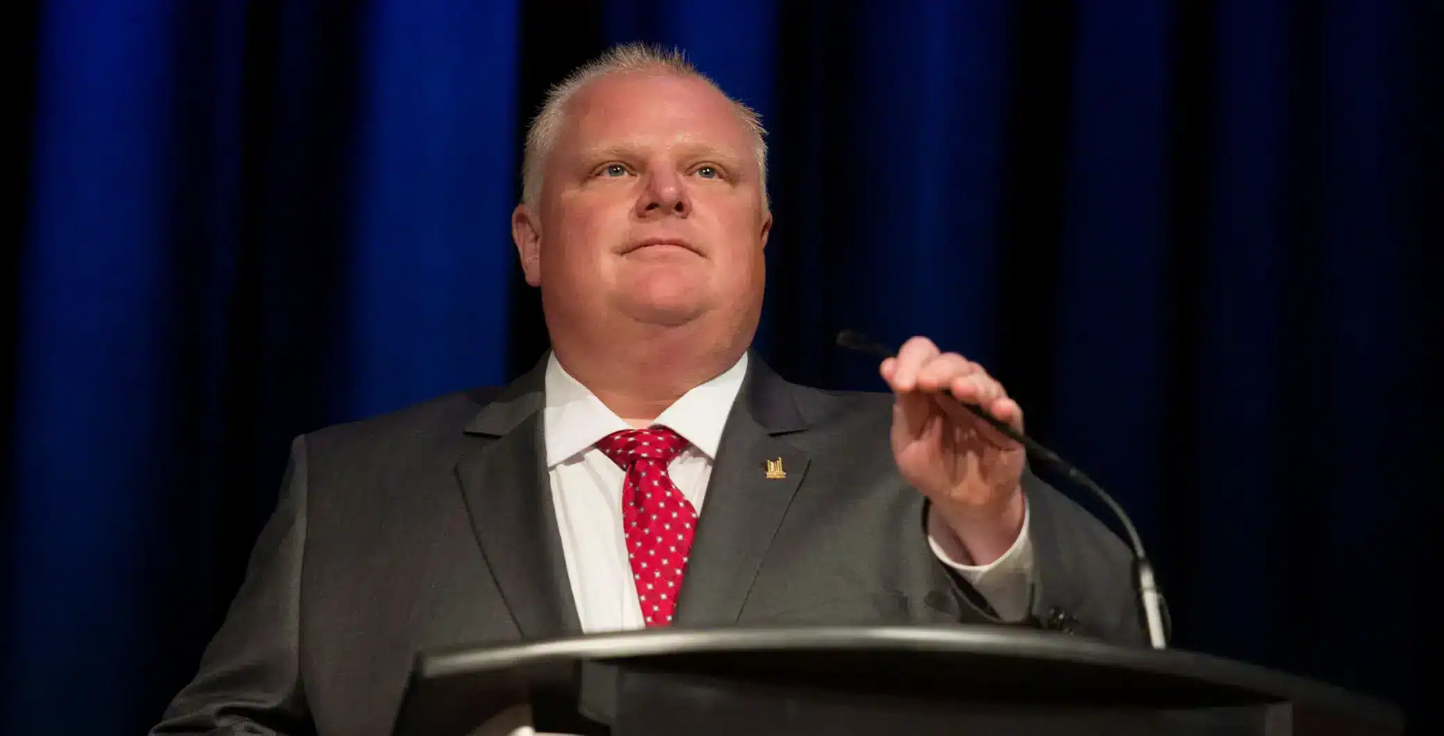 Professional male speaker at a podium during a formal event, wearing a suit and red patterned tie, with a dark blue curtain backdrop, delivering a speech or presentation in Toronto.