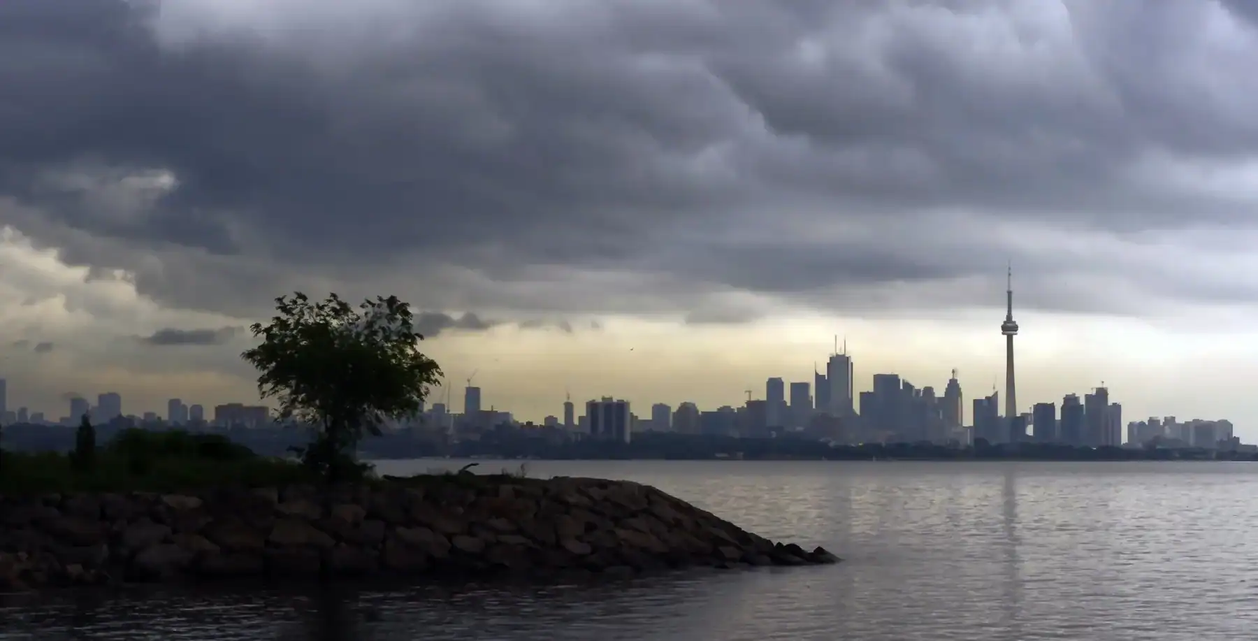 City skyline of Toronto with CN Tower and cloudy sky over waterfront, urban landscape, modern skyscrapers, downtown Toronto scenic view, Canada cityscape, water reflection, moody weather.