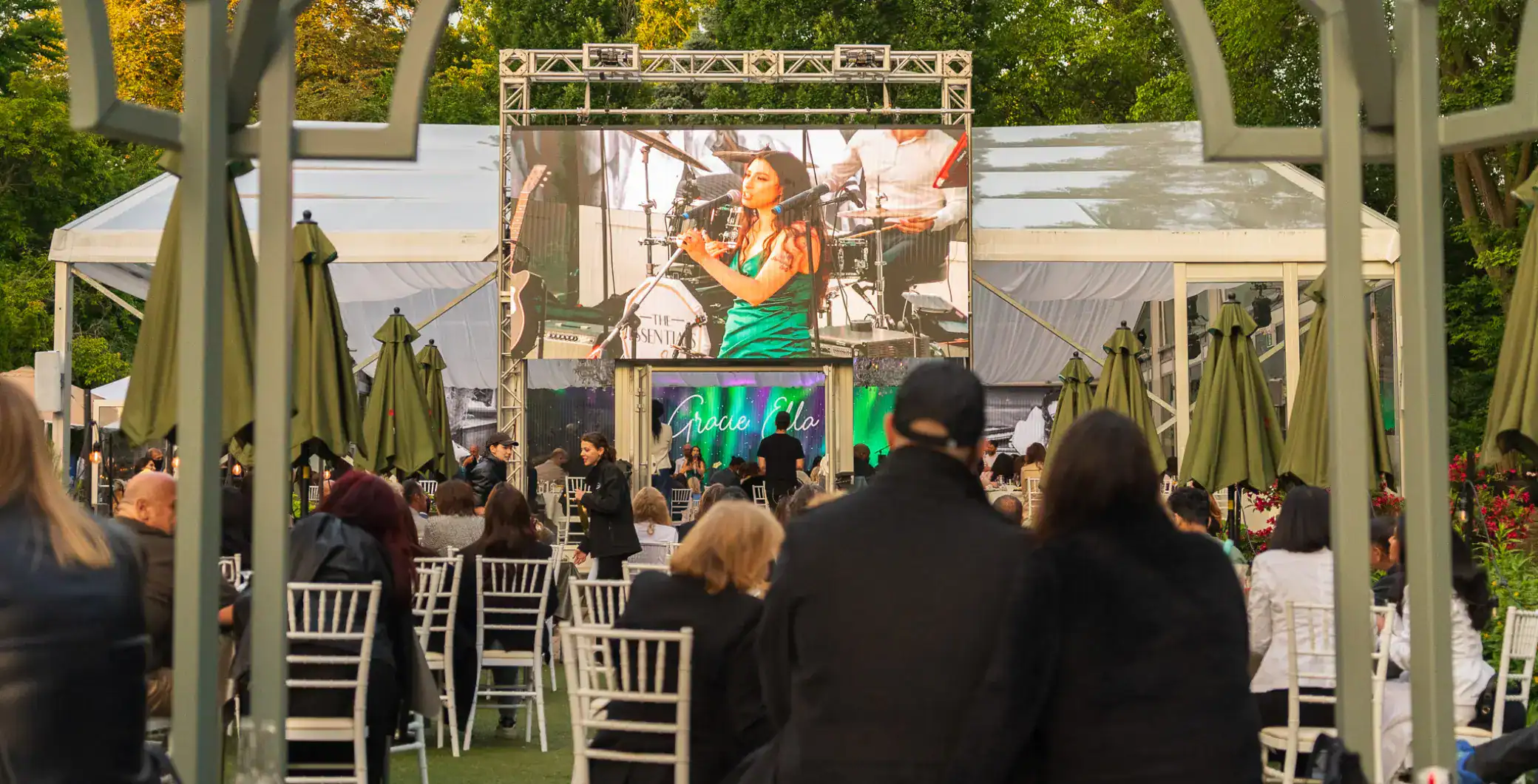 Vibrant outdoor concert at NOW Toronto festival with live music, large screen, and audience seated on lawn under green umbrellas in Toronto park.