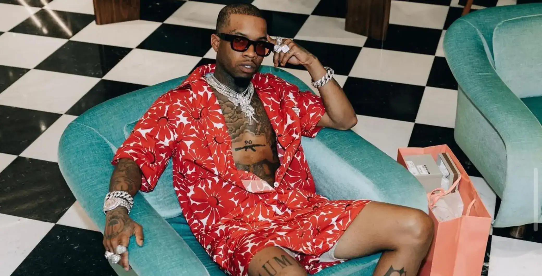 Luxury rapper with tattoos wearing red floral shirt and sunglasses lounging on modern teal sofa in upscale Toronto setting.