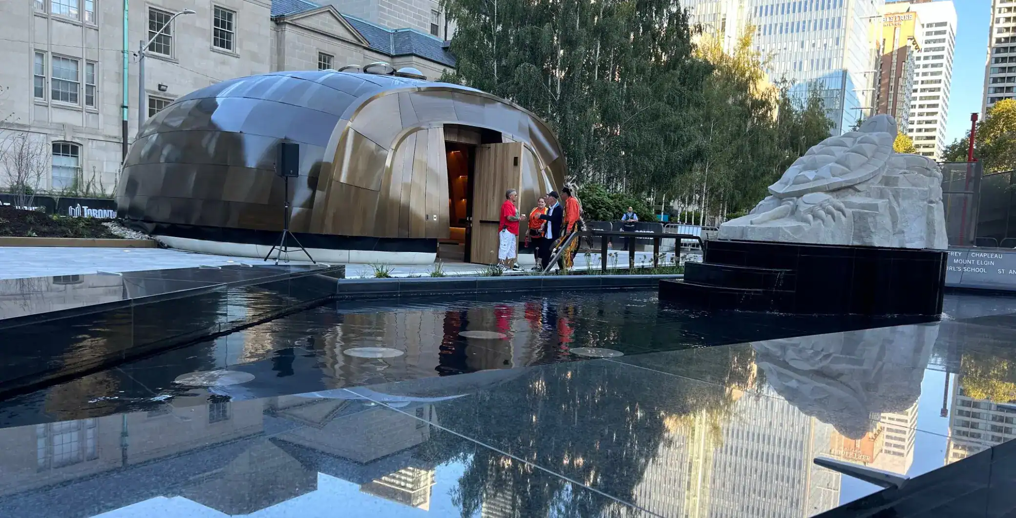 Futuristic architectural structure with a reflective water feature in an urban setting, surrounded by tall buildings and cityscape, highlighting Toronto downtown attractions and modern design.