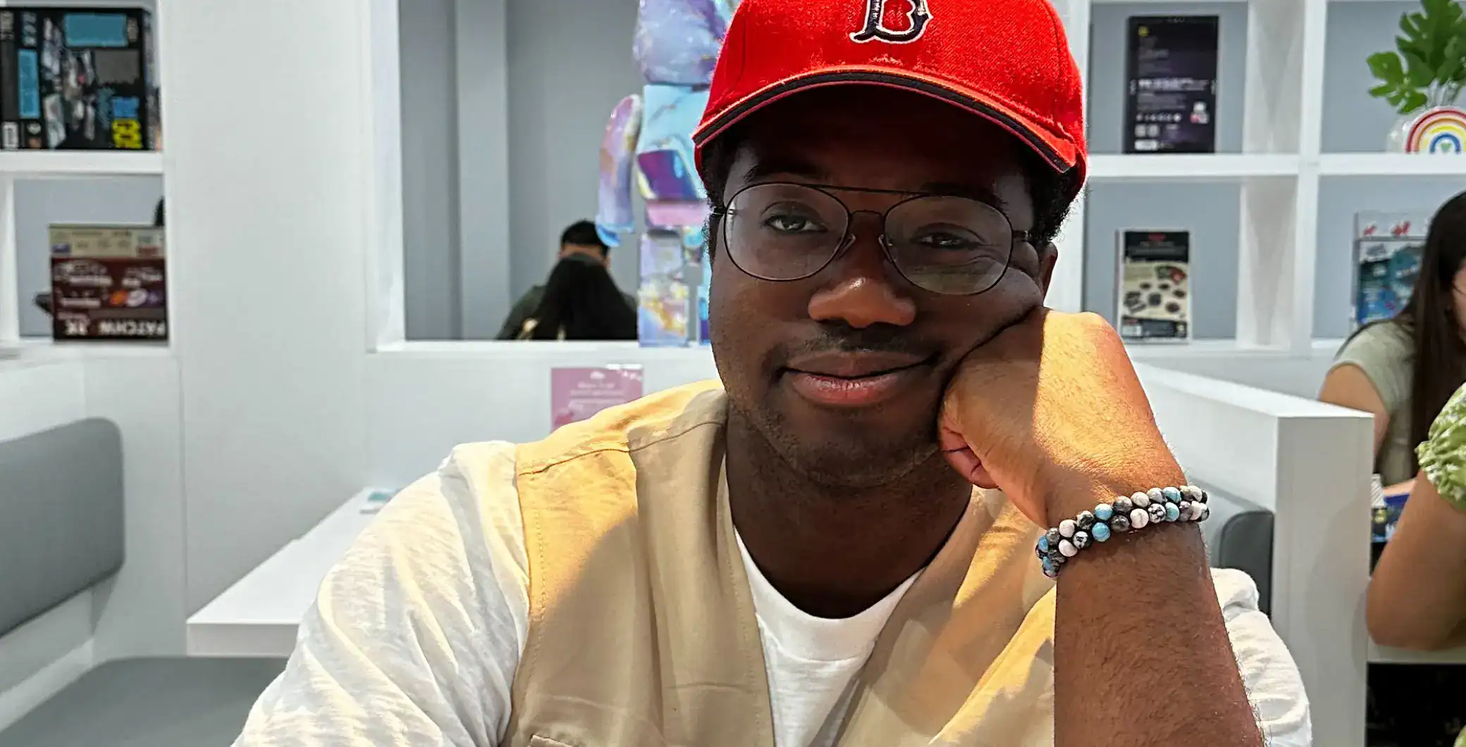 Bright young man smiling at a cafe in Toronto, wearing a red cap, glasses, and a beaded bracelet, enjoying a casual day out in the city.