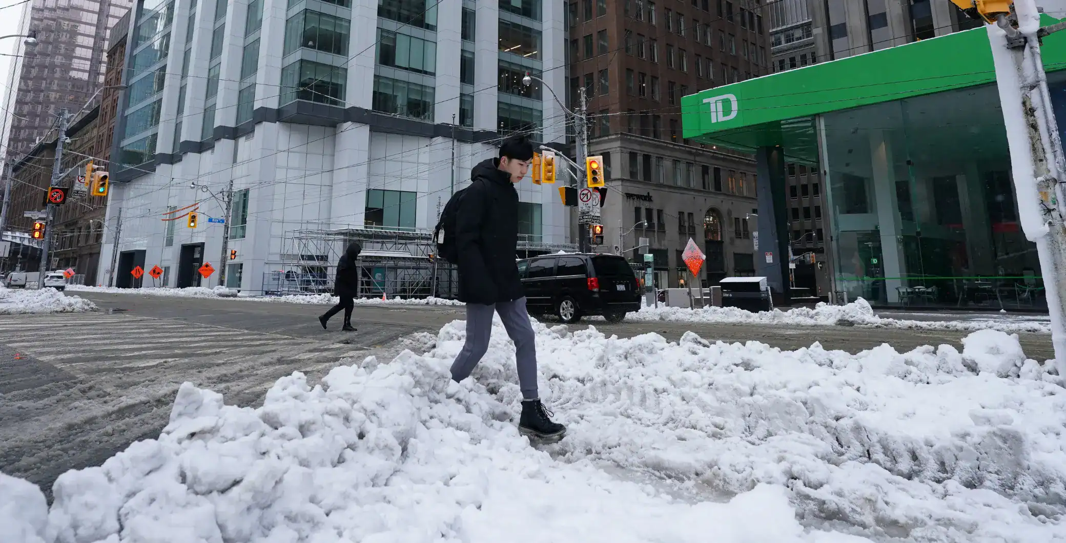 Young man walking through snow on Toronto city street with tall modern buildings and TD Bank branch in background, winter in downtown Toronto, urban winter scene.