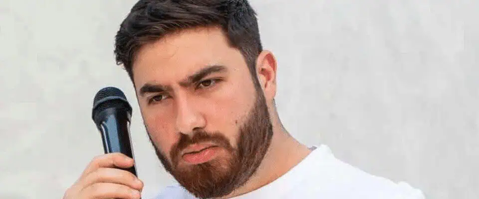 Young man with a beard holding a microphone, performing or speaking on stage.