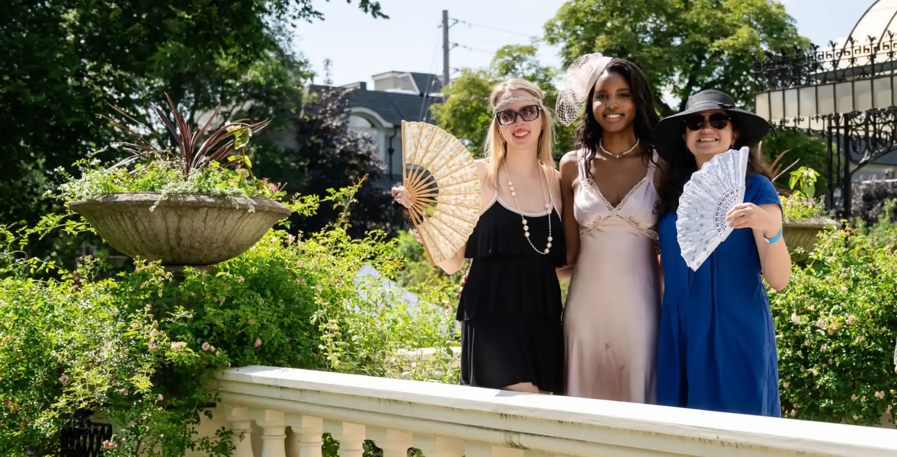Elegant women in vintage dresses holding lace fans outdoors in Toronto garden, celebrating spring or summer, with lush greenery and historic architecture in the background.