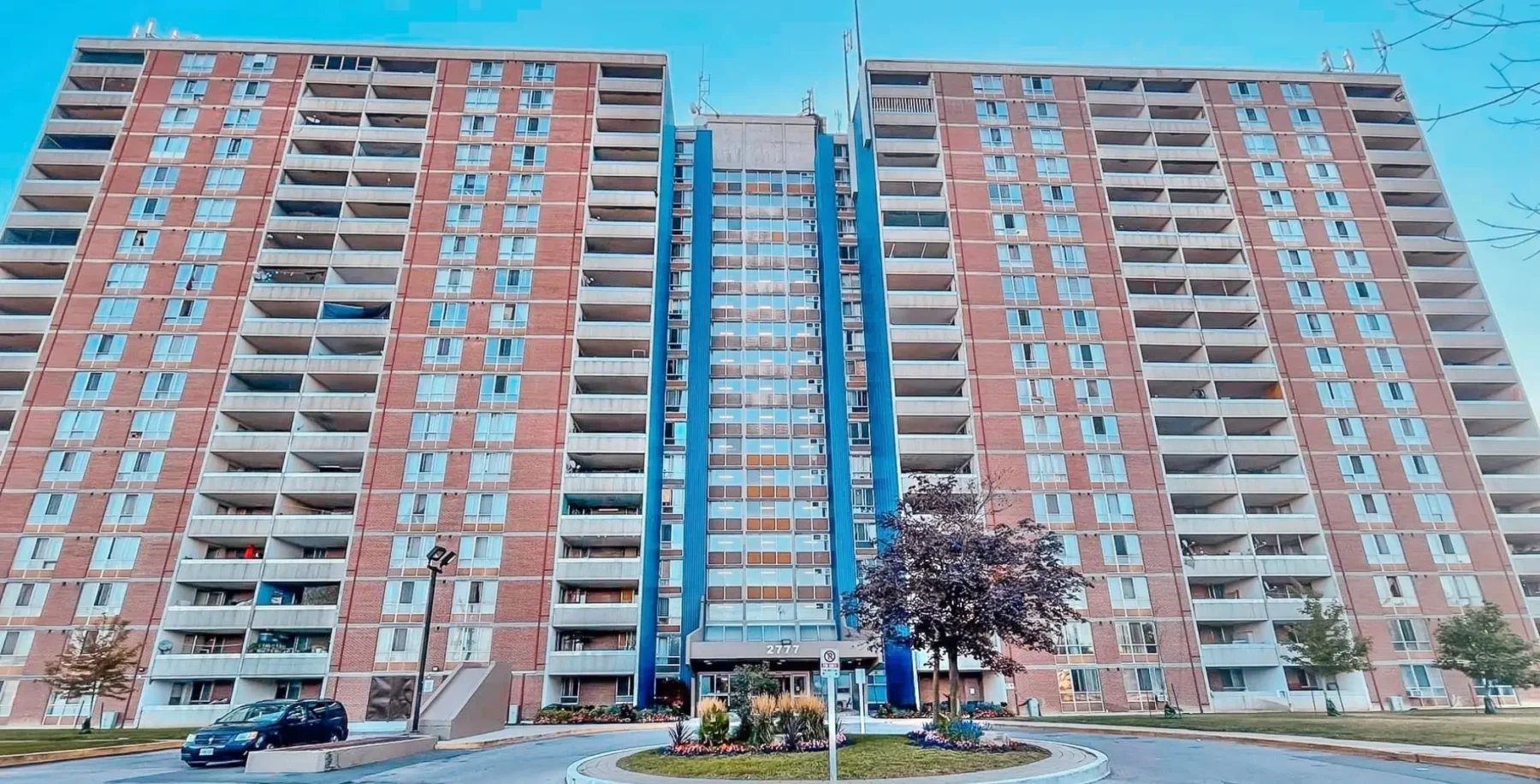 Large Toronto apartment building with multiple units, blue central elevator shaft, and parking lot in front, illustrating urban residential living.
