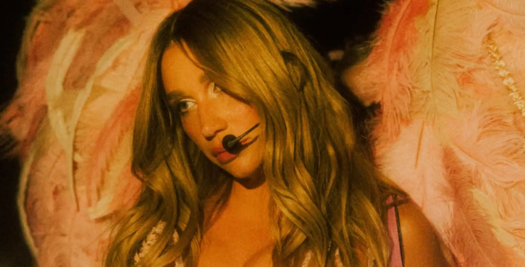 A young woman with long wavy hair wearing a headset microphone, performing on stage with pink feathered wings in the background, at a live event or concert.
