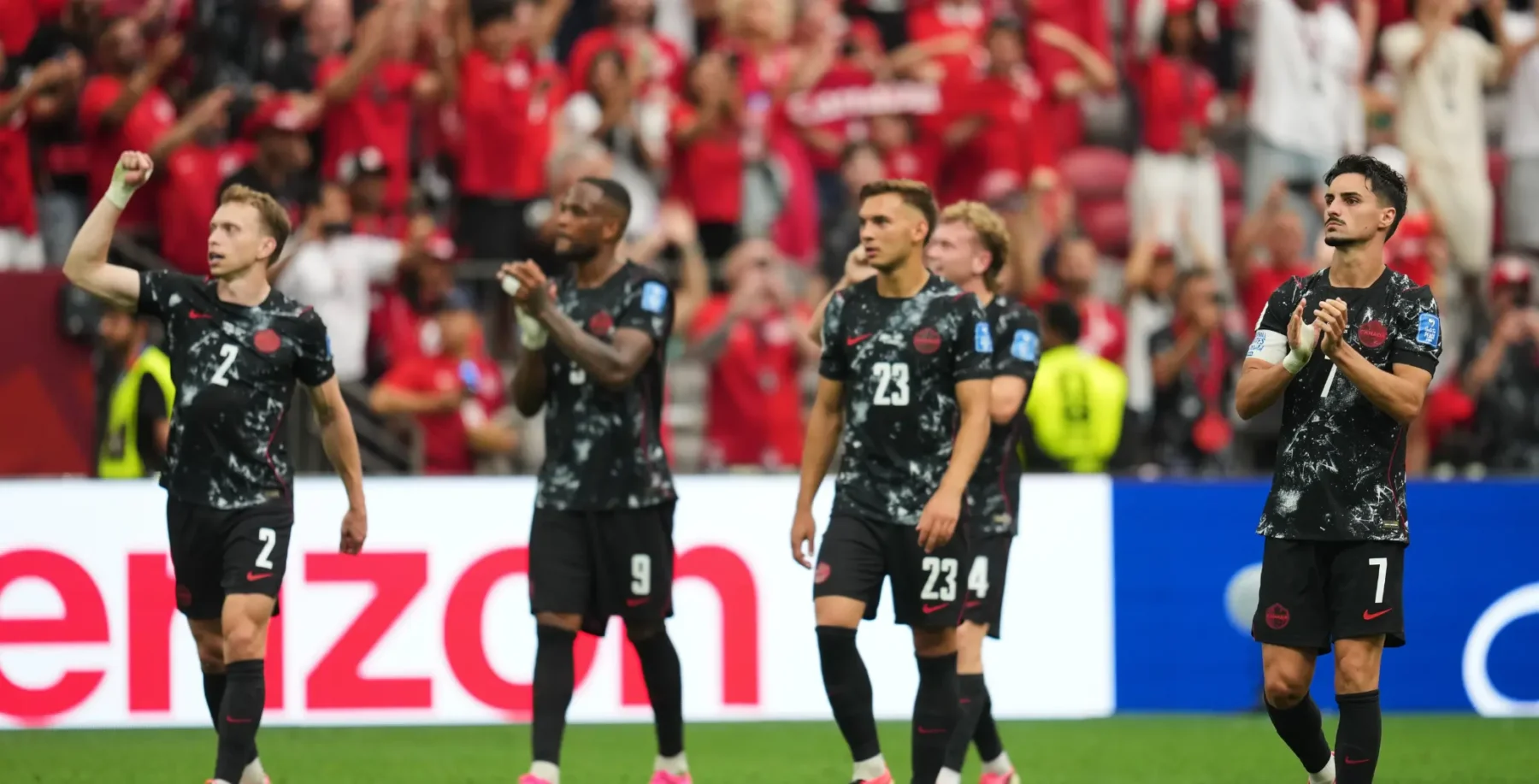 Canadian soccer players celebrating on the field during a match with fans in the background.