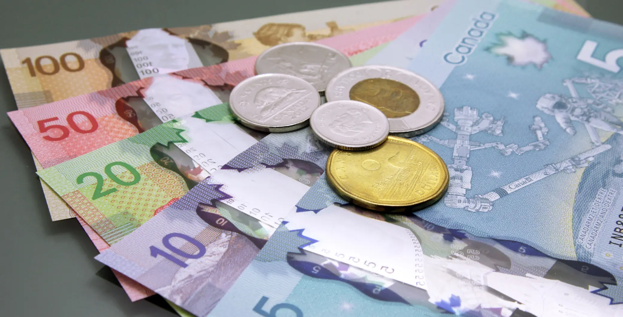 A colourful assortment of Canadian banknotes and coins, including denominations of 5, 10, 20, 50, and 100 dollars, spread out on a flat surface, representing Canadian currency.