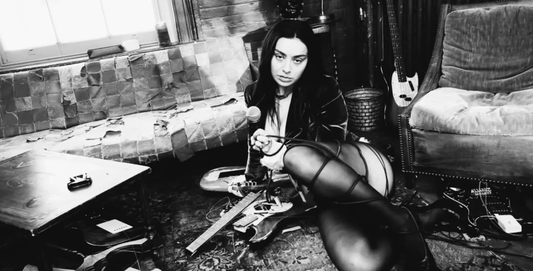 A young woman with dark hair and bold makeup sitting on the floor of a music studio, surrounded by musical instruments and equipment, in a black-and-white photo.