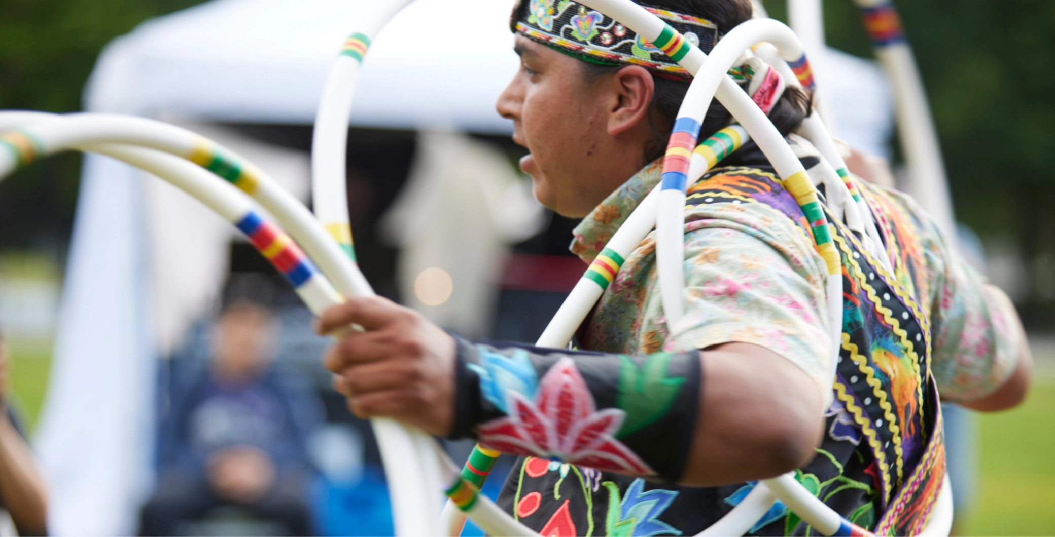 A woman wearing a colorful, patterned outfit and headband, holding white hoops during a cultural outdoor event in Toronto, showcasing traditional dance and vibrant community celebration.