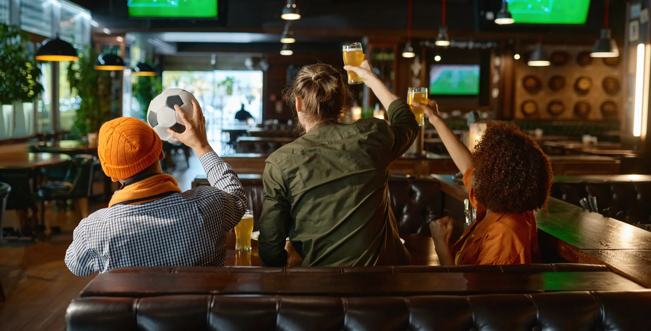 Three friends watch a sports game on TV at a bar, cheering and holding drinks, with a lively atmosphere and multiple screens in the background.