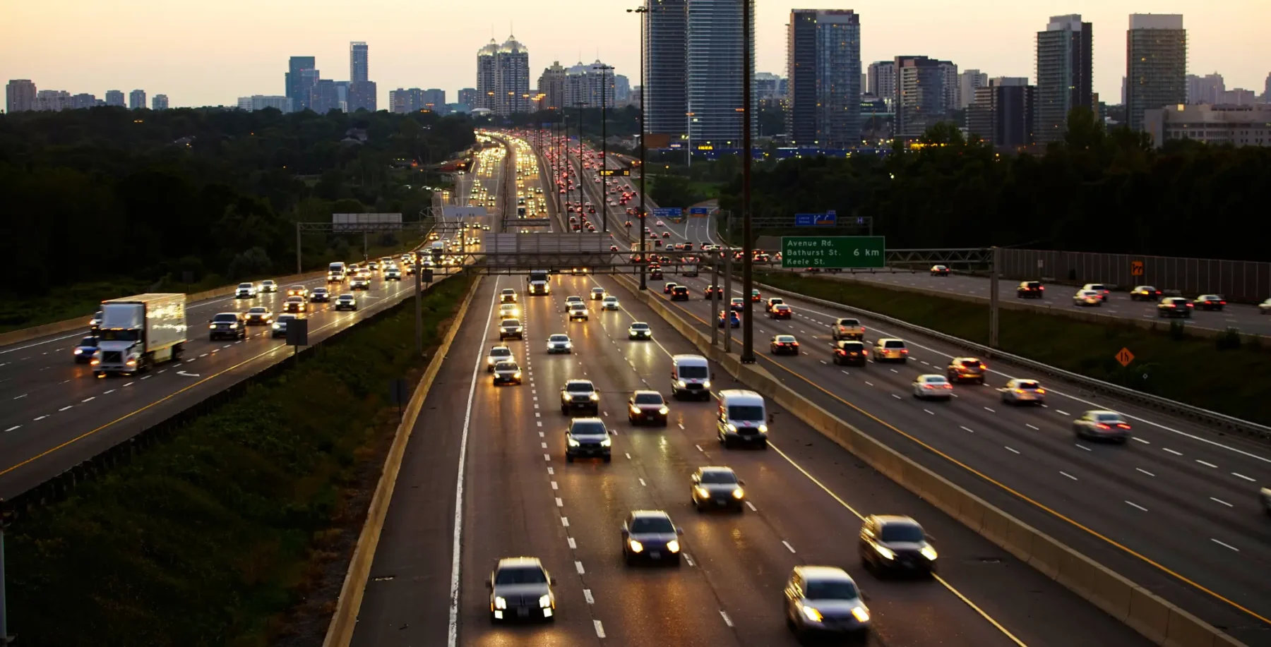 Toronto highway with multiple lanes of traffic during evening rush hour, city skyline in the background.