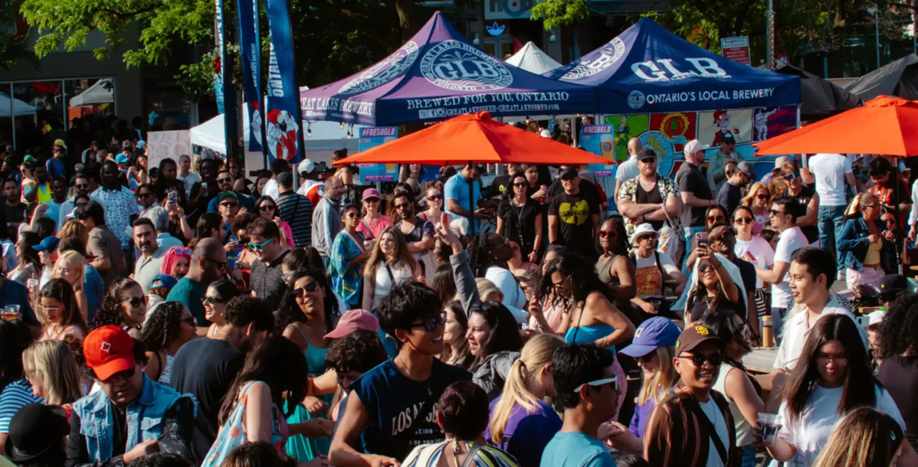 A lively outdoor event in Toronto with a large crowd, colorful tents, and orange umbrellas, showcasing community gathering, local breweries, and city entertainment in a vibrant urban setting.