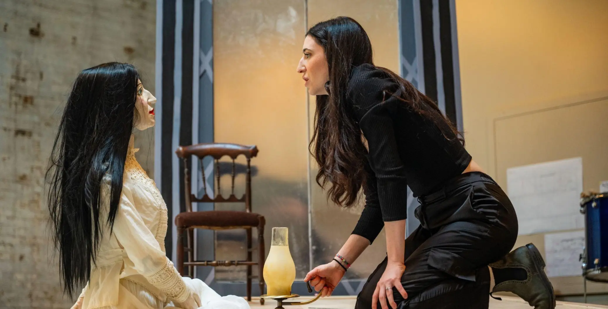 A young woman with long dark hair, dressed in black, leaning forward and speaking intensely to a seated woman with long black hair, wearing a cream-colored sweater, in an indoor setting.
