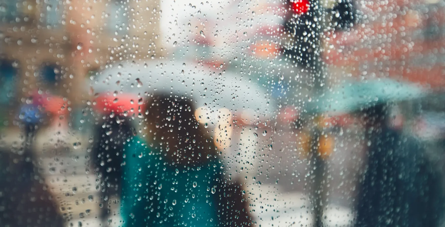 People with umbrellas walking in the rain on a city street through a rain-covered window.