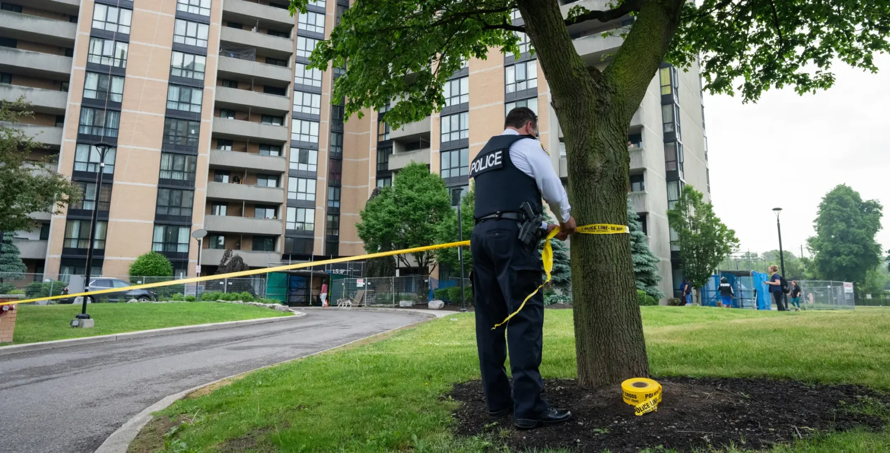 A police officer securing a tree in a park with yellow caution tape near residential buildings in Toronto, Ontario, during daytime.