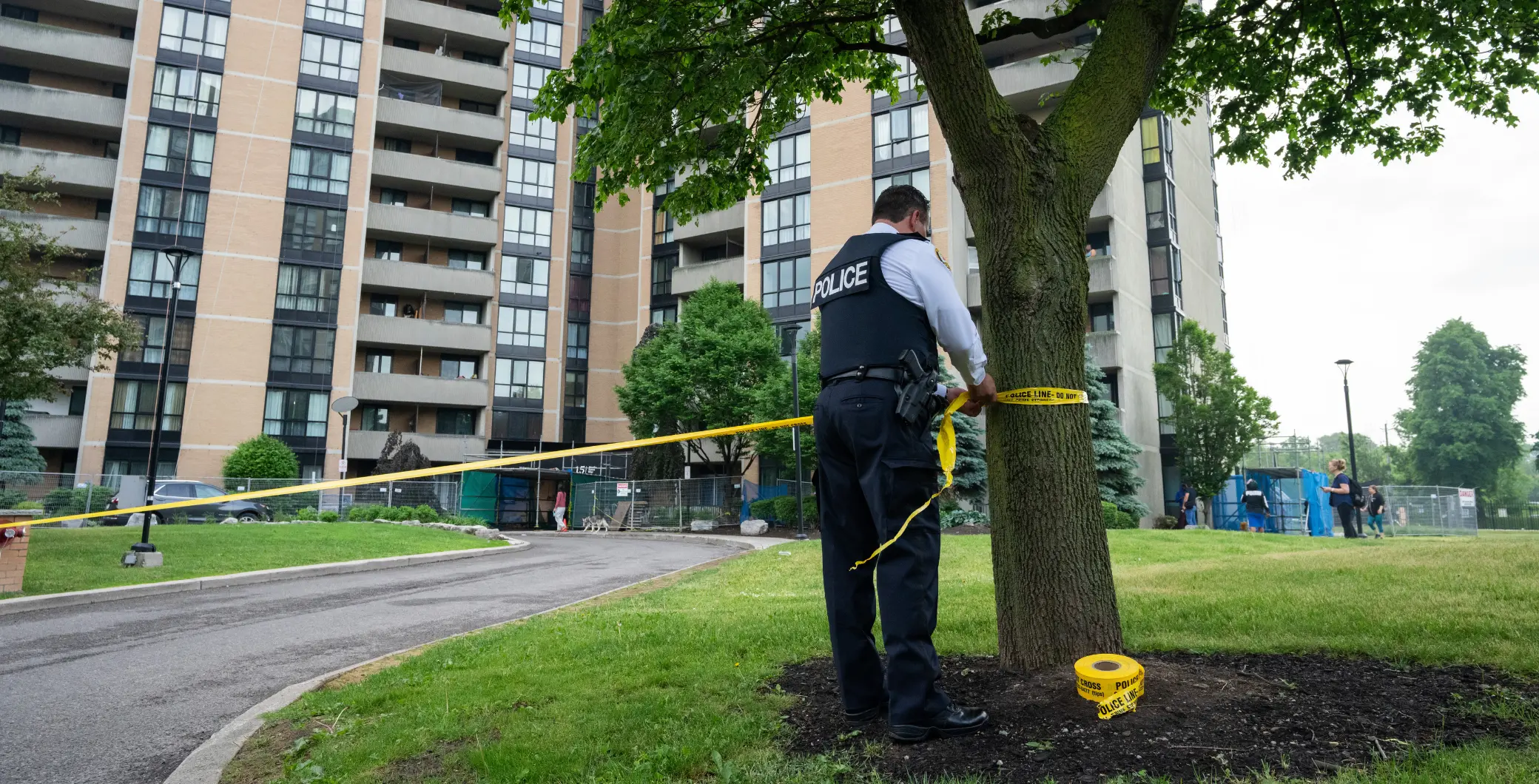 A police officer securing a tree in a park with yellow caution tape near residential buildings in Toronto, Ontario, during daytime.