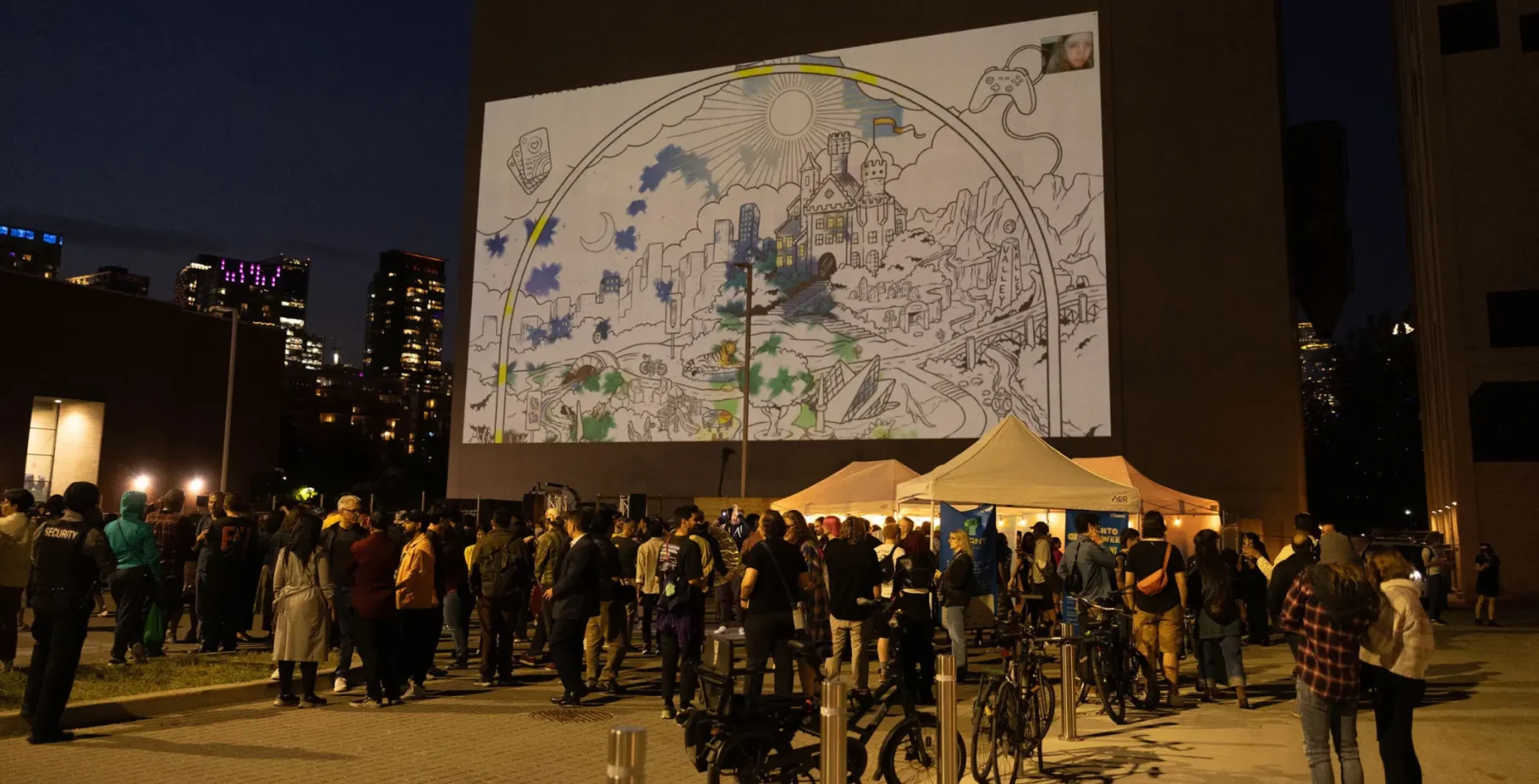 A vibrant outdoor event in Toronto featuring a large illuminated mural projected on a building, with a crowd gathered around tents and bicycles, showcasing local arts and community engagement.