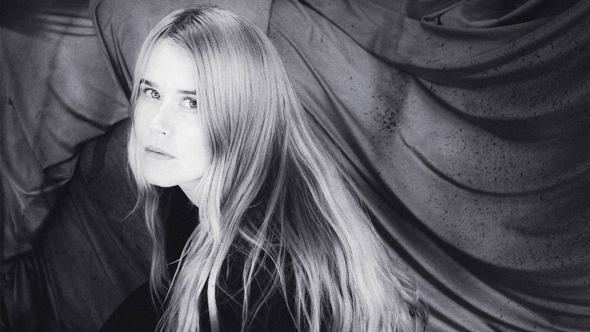 Black and white portrait of a woman with long blonde hair, looking at the camera with a neutral expression, against a textured fabric backdrop.