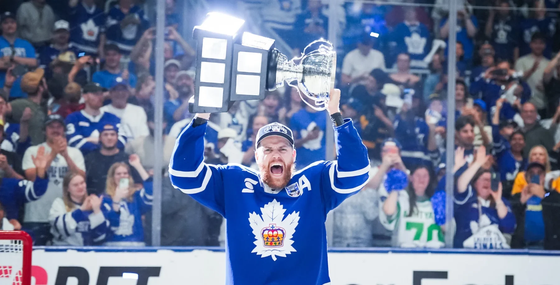 Hockey player in blue jersey celebrating with trophy after winning game, surrounded by cheering fans in arena.
