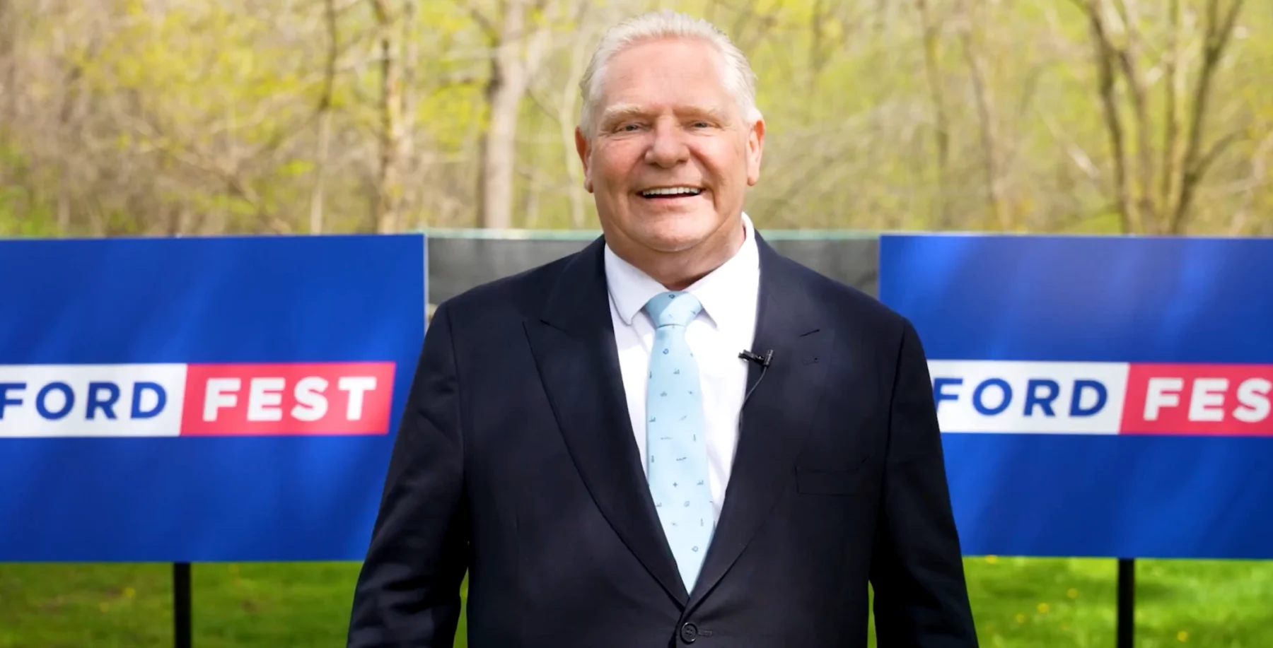 A smiling man in a suit and tie standing outdoors at a Ford Fest event with blue banners in the background, promoting community engagement and local celebrations in Toronto.