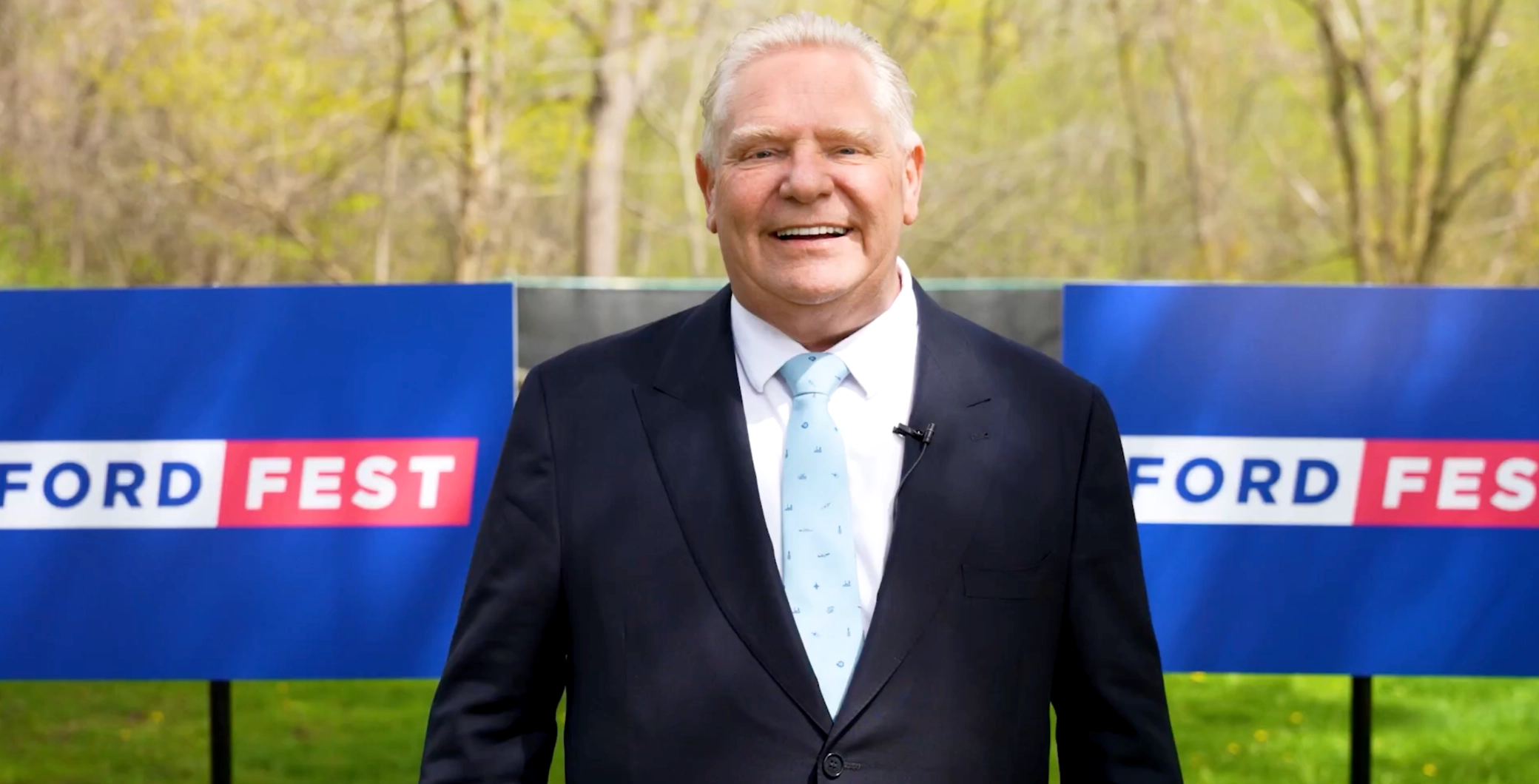 A smiling man in a suit and tie standing outdoors at a Ford Fest event with blue banners in the background, promoting community engagement and local celebrations in Toronto.