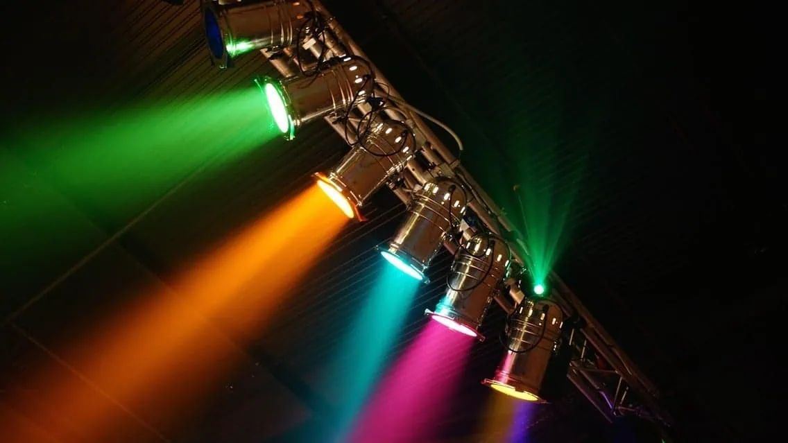 Stage lighting with colorful spotlights in green, orange, blue, pink, and yellow illuminating a dark performance space.