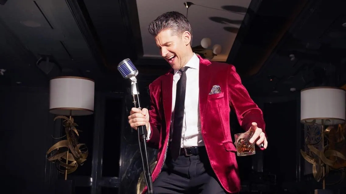 Matt Dusk performing on stage in a red velvet blazer, holding a microphone and a glass of drink, during a Christmas-themed concert.