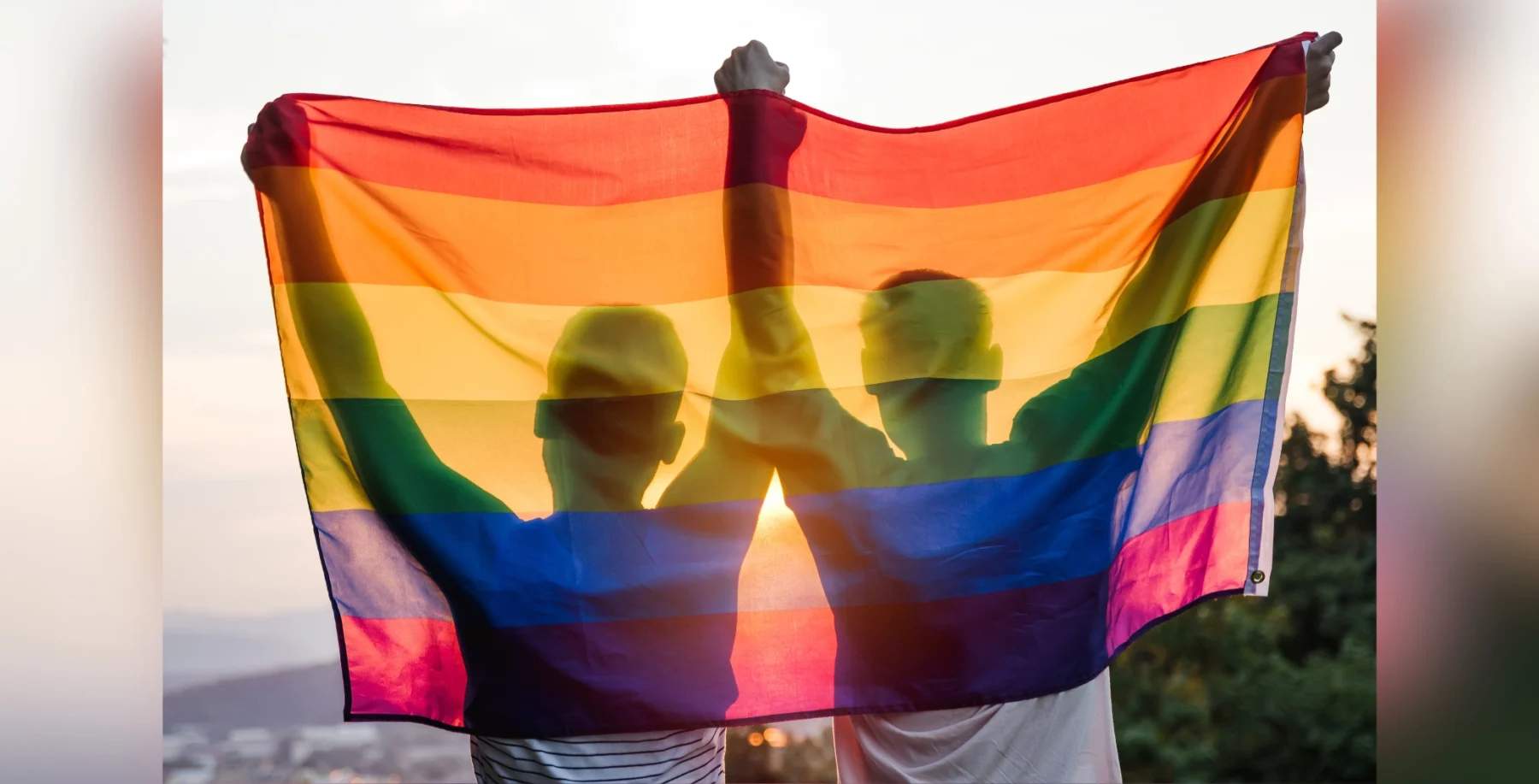 Two people holding a rainbow pride flag outdoors at sunset, symbolizing LGBTQ+ pride and support.