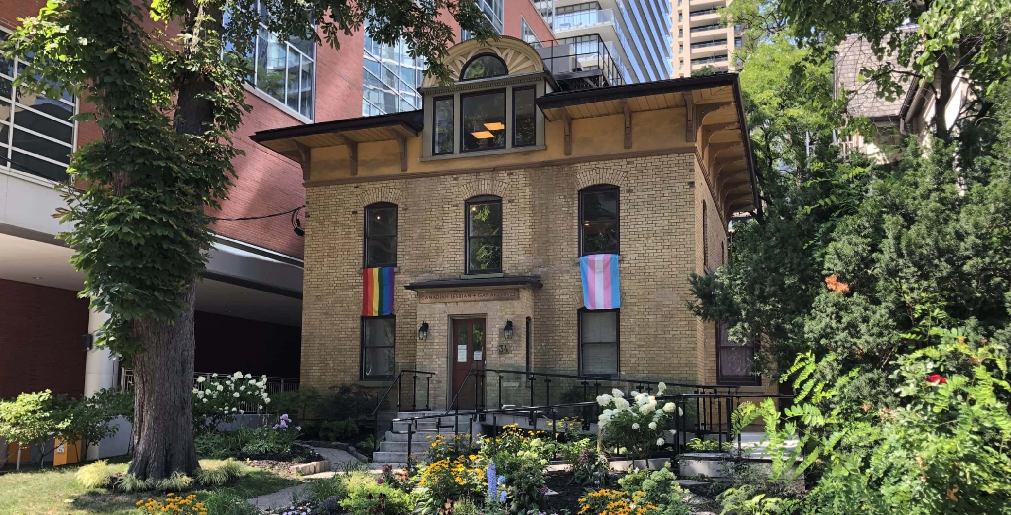 A historic brick house surrounded by lush greenery and colorful flowers in an urban setting, representing Toronto's vibrant neighborhood and architectural diversity.