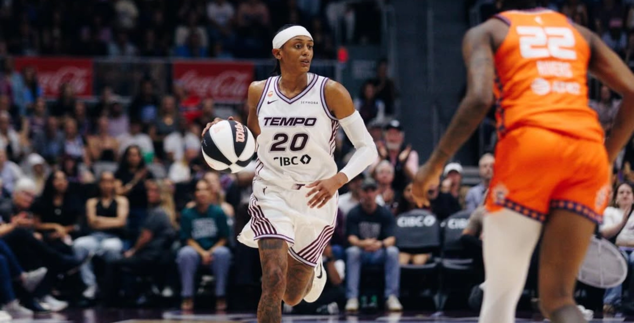 A woman basketball player in a white uniform with the number 20 dribbling the ball during a game at an indoor arena with spectators.
