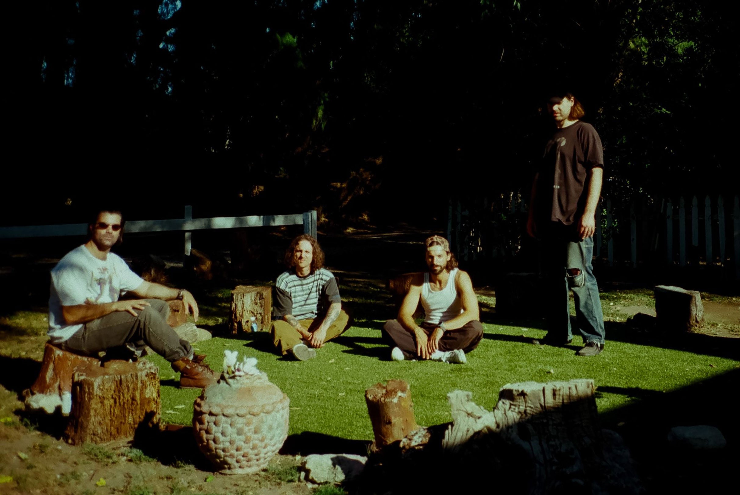 Four people sitting and standing outdoors on grass with trees in the background, in a casual setting for a music tour promotion.
