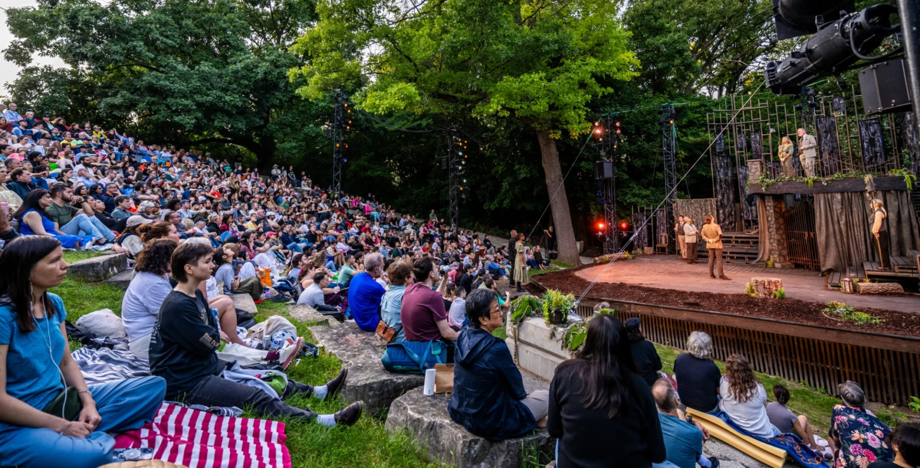Audience watching an outdoor Shakespeare play in a park with a large crowd seated on the grass and trees in the background.