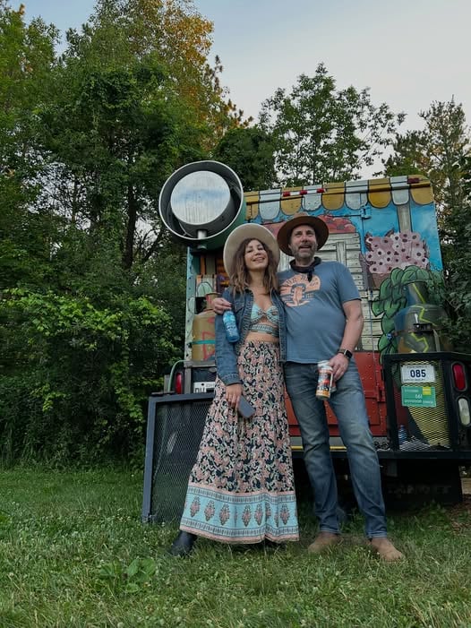 Two people stand in front of a colorful food truck with trees in the background, smiling and enjoying a sunny day.