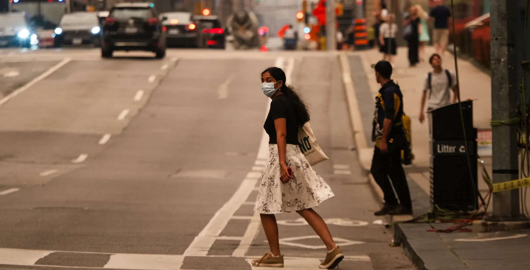 Woman crossing Toronto street wearing a face mask during evening rush hour with cars and pedestrians in the background.